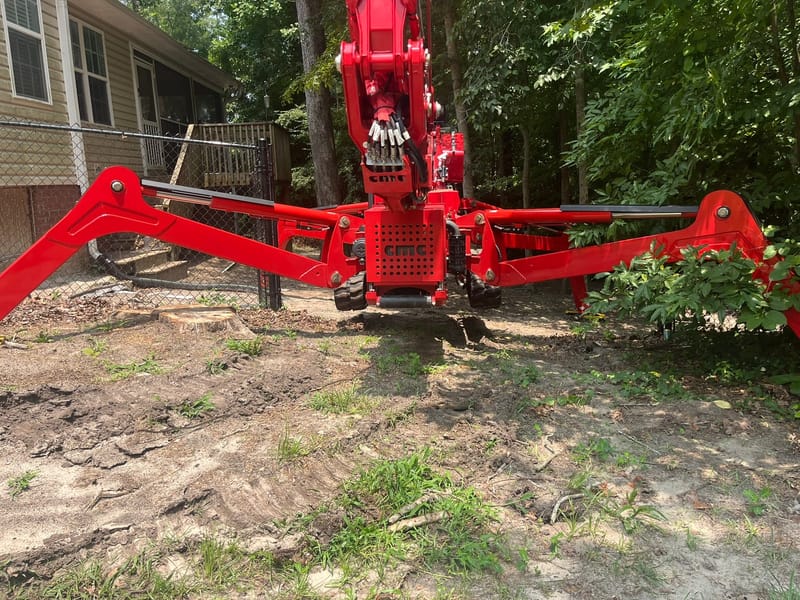 Red tracked lift set down on its four outriggers on bare ground tight against the side of a house.